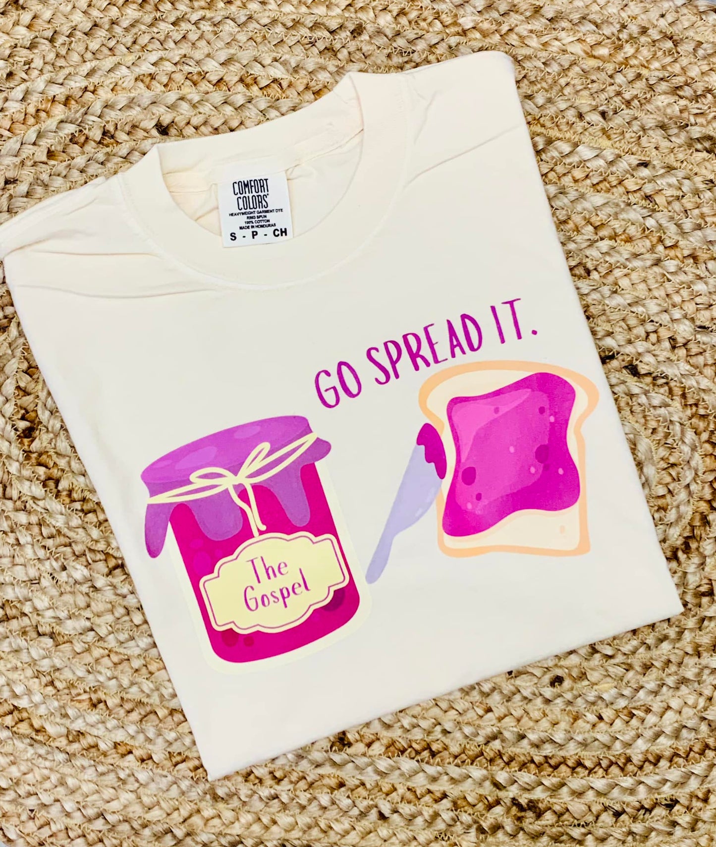 A youth-sized white t-shirt with a graphic print of a jar and a slice of bread, alongside the phrase 'GO SPREAD IT.'
