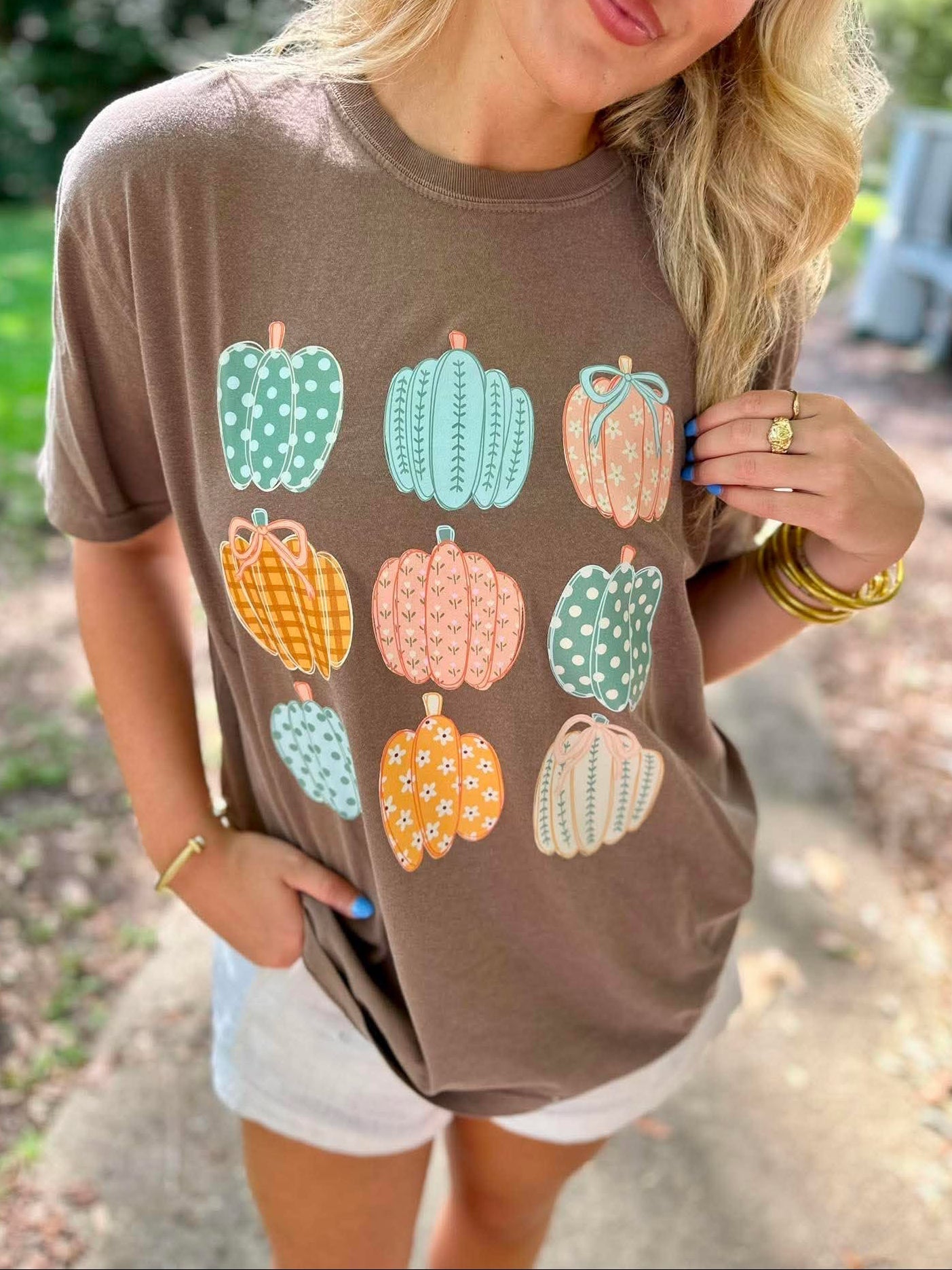 A woman wearing a brown short-sleeve t-shirt with a graphic print of colorful patterned pumpkins, photographed outdoors on a path.