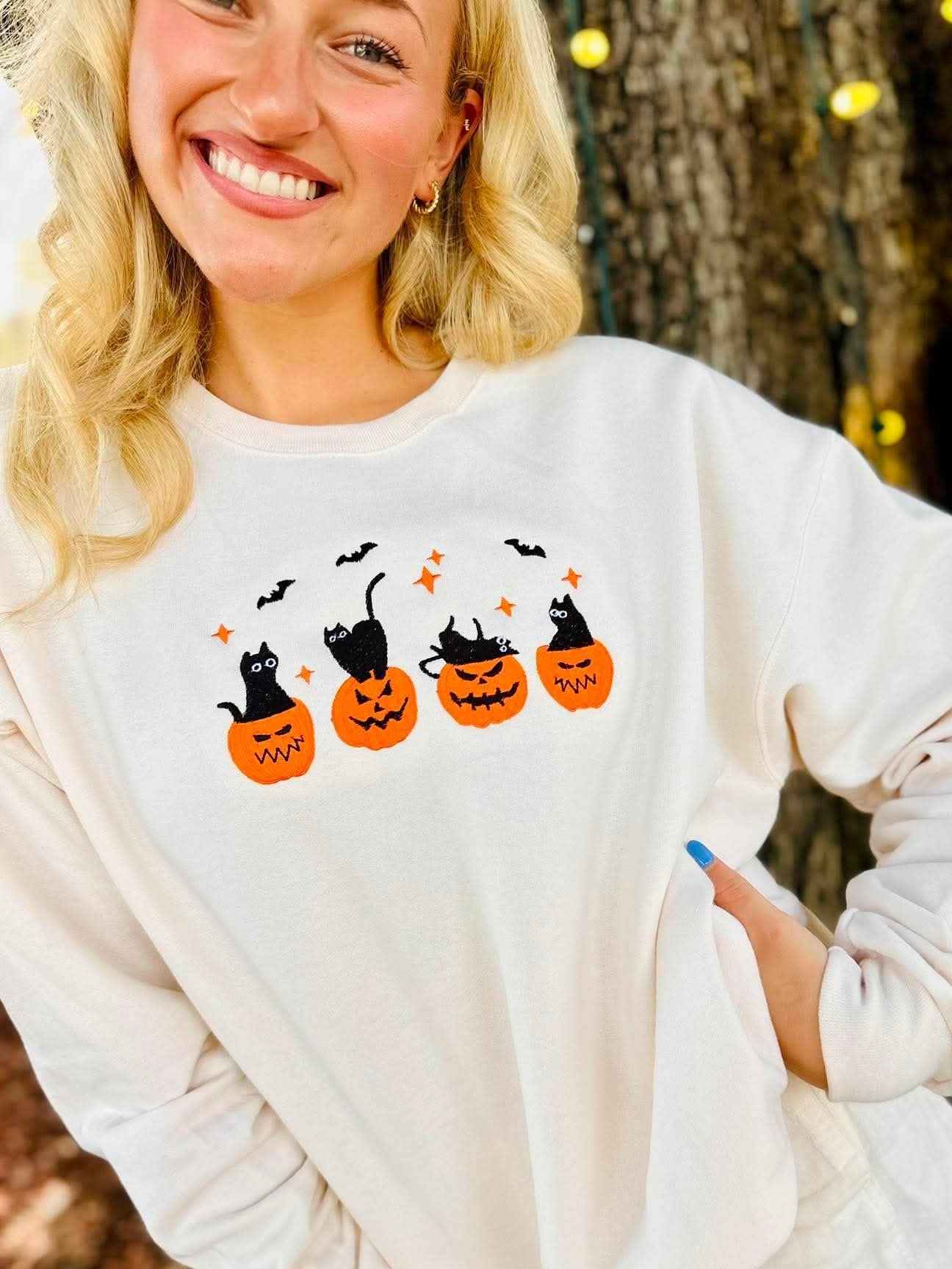 A smiling blonde woman wearing a white long-sleeve sweatshirt with a Halloween graphic of three black cats in orange pumpkins, posed outdoors in front of a tree trunk with warm string lights.