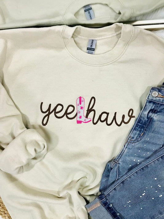 A tan embroidered sweatshirt with the text 'YEE HAW' on it, displayed on a surface with blue jeans and other clothing items.