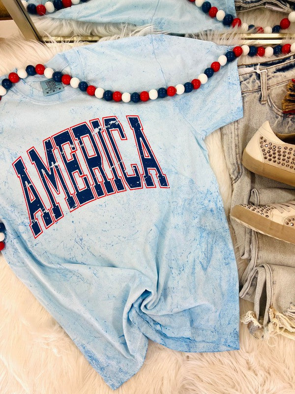A blue colored graphic tee with the text 'AMERICA' printed in red and white, displayed on a faux fur surface with jeans and a decorative lei in the background.