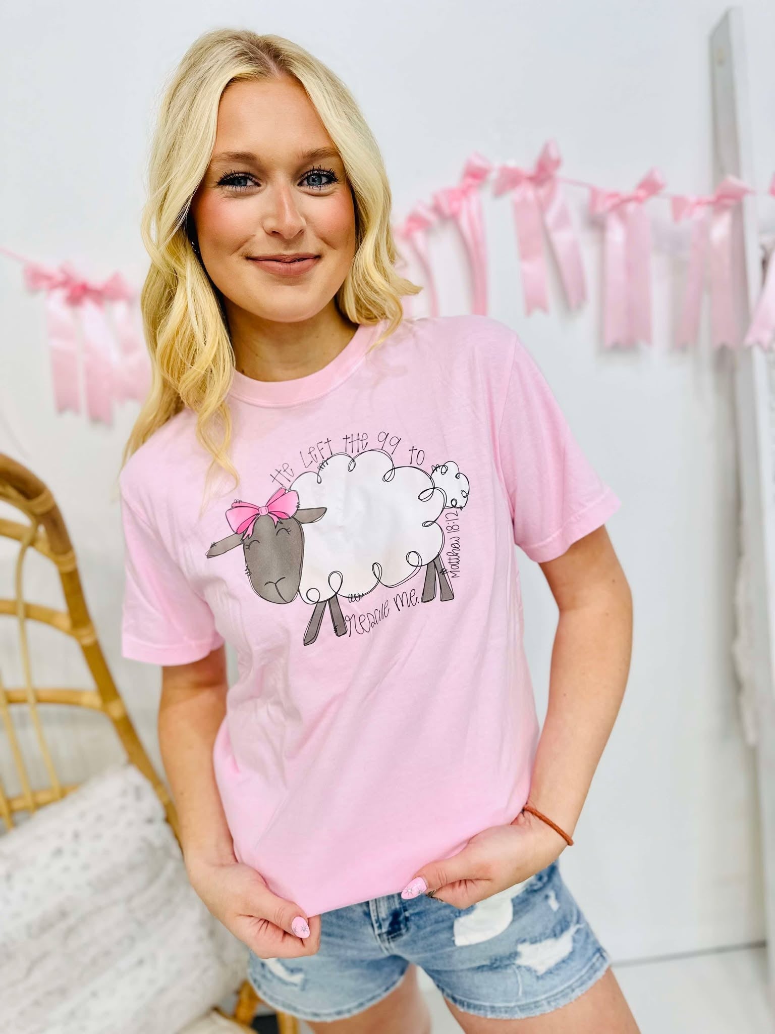 A person wearing a pink graphic tee with text and a sheep design, standing in front of a white background with decorative elements.