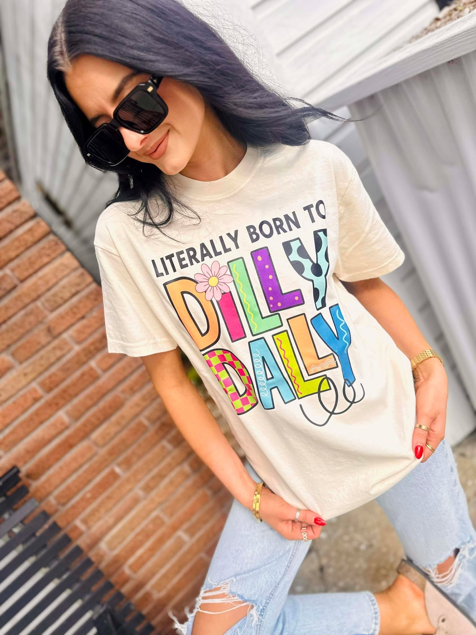 Woman wearing a Born to Dilly Dally Ivory  t-shirt with colorful text outdoors