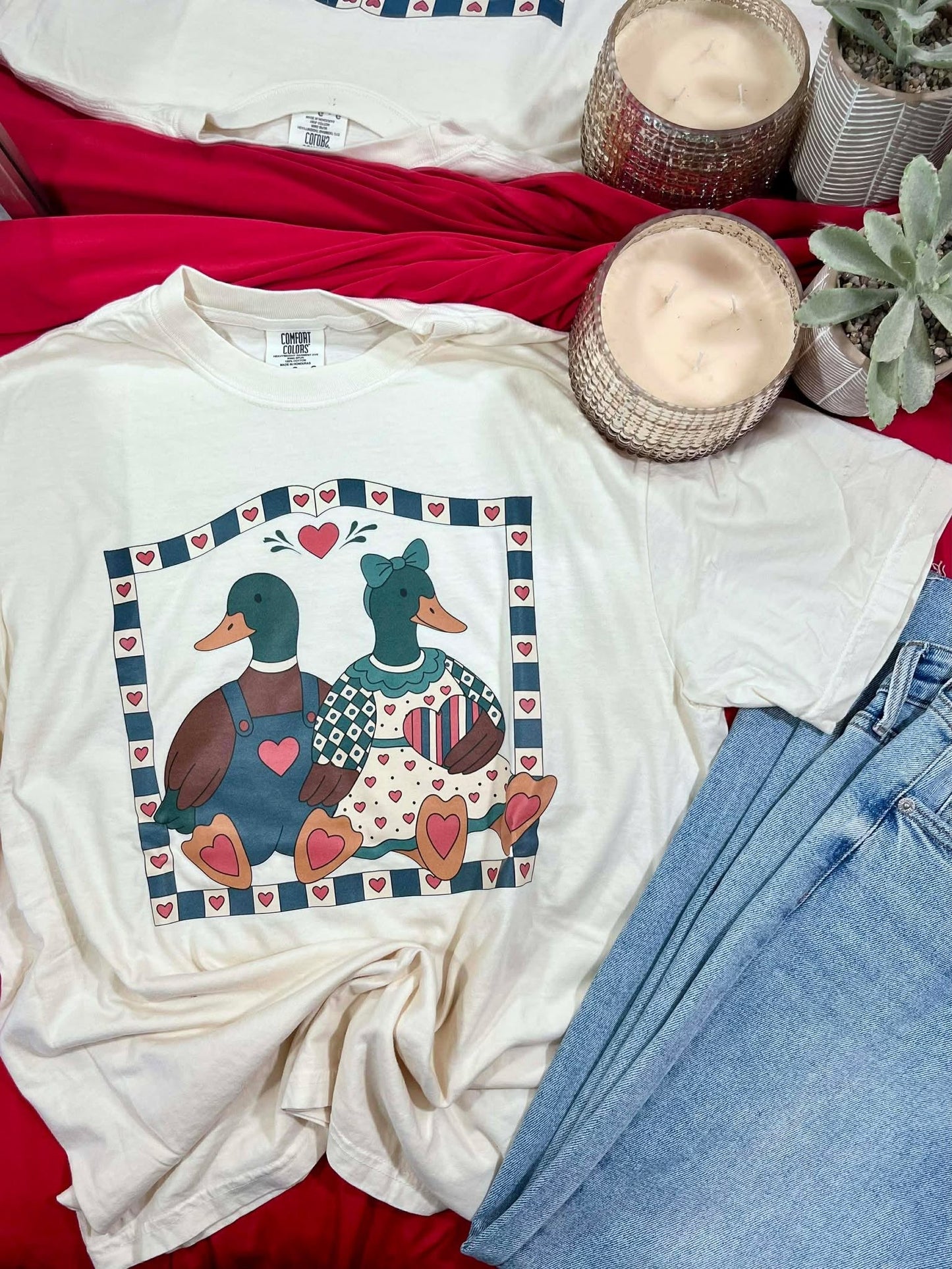 White t-shirt with duck design on a red surface with jeans and candles.