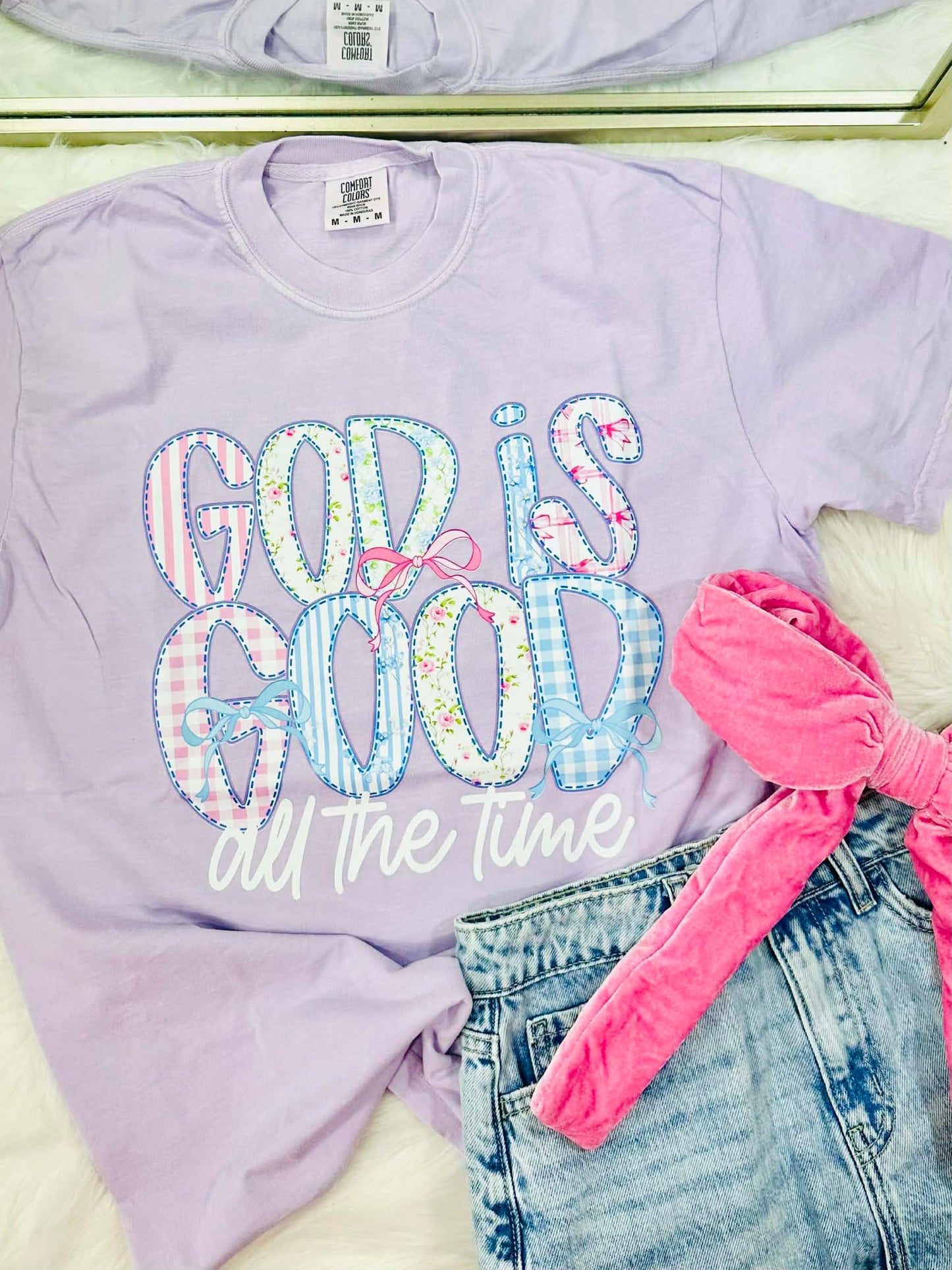 Lavender sweatshirt with 'God is good all the time' text, pink bow, and denim shorts.