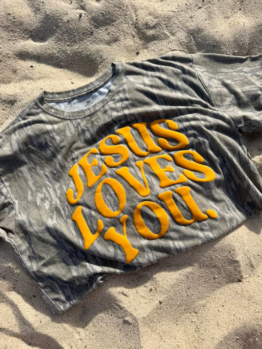 Gray t-shirt with 'Jesus Loves You' in yellow on a sandy surface