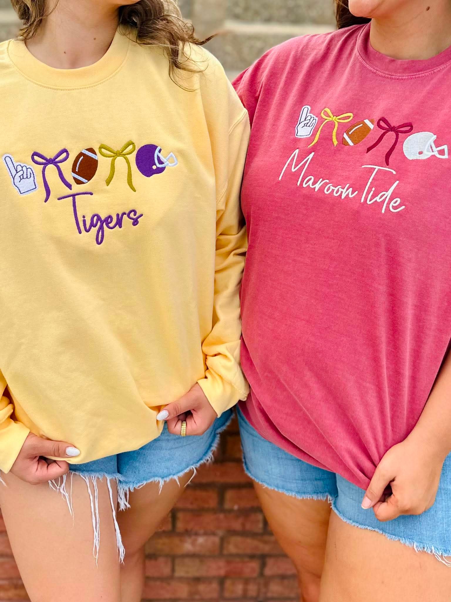 Two women wearing graphic crewneck sweatshirts, one in pale yellow with purple and gold football-themed graphics and the word 'Tigers', and the other in heather pink with white 'Maroon Tide' script and red, gold, and white graphics. They are wearing denim shorts against a brick wall background.