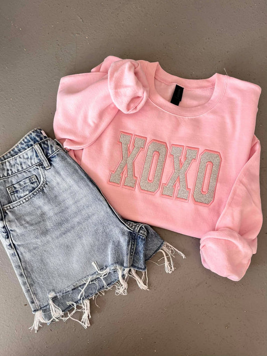 Pink sweatshirt with 'XOXO' Glitter Applique embroidery and denim shorts on a gray background