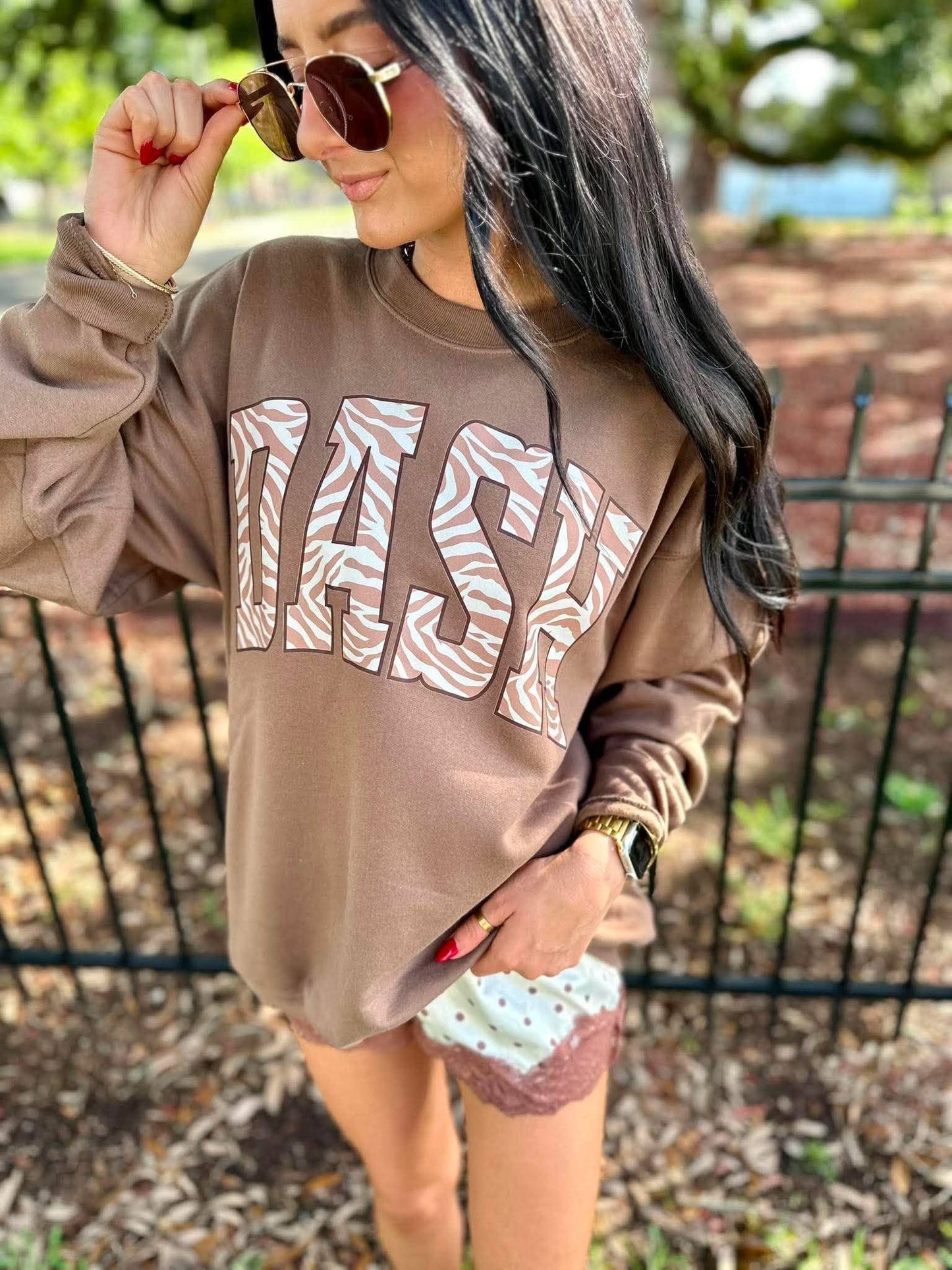 A woman wearing a brown sweatshirt with 'DASH' printed in large white and tan zebra-patterned letters, paired with patterned shorts and sunglasses, standing outdoors in front of a fence.