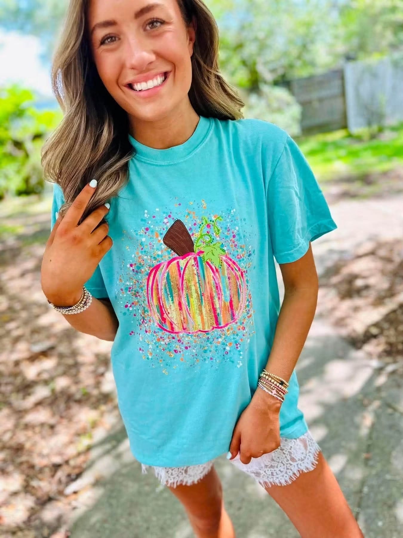 A woman wearing a turquoise graphic t-shirt with a colorful painted pumpkin design, paired with white lace-up shorts, standing outdoors on a paved path with green foliage in the background.