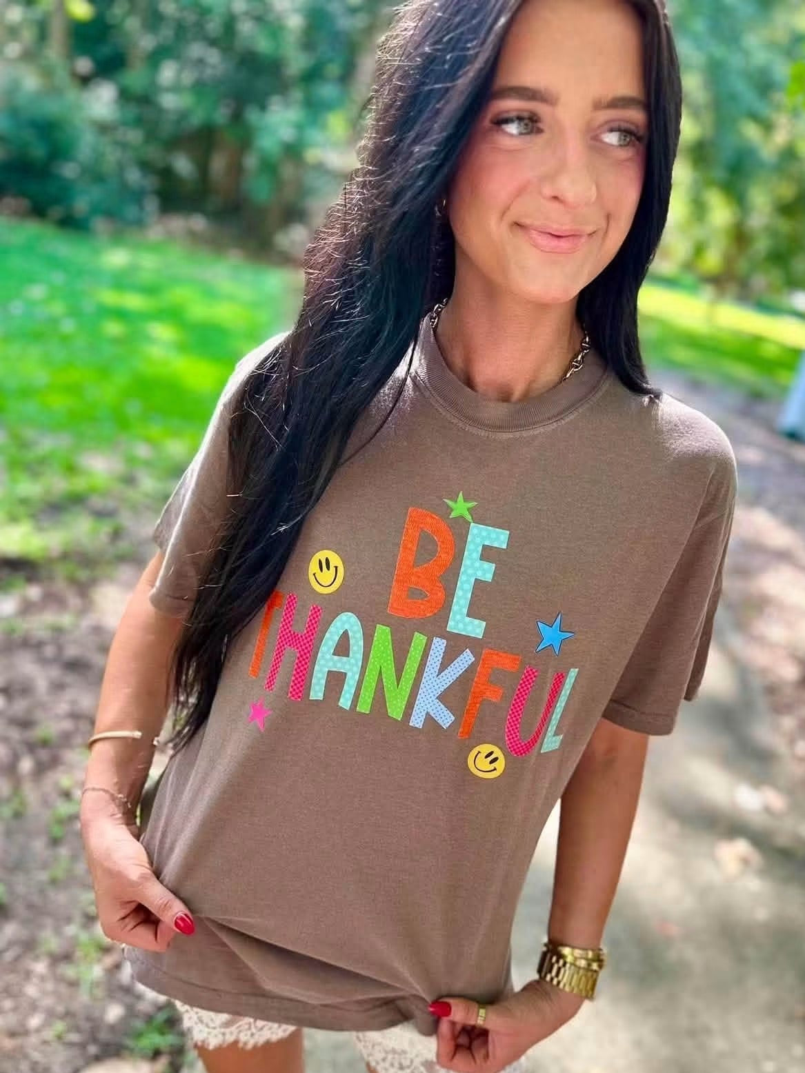 A woman wearing a brown graphic t-shirt with the word 'BE THANKFUL' in colorful letters and smiley faces, standing outdoors on a path with trees in the background.