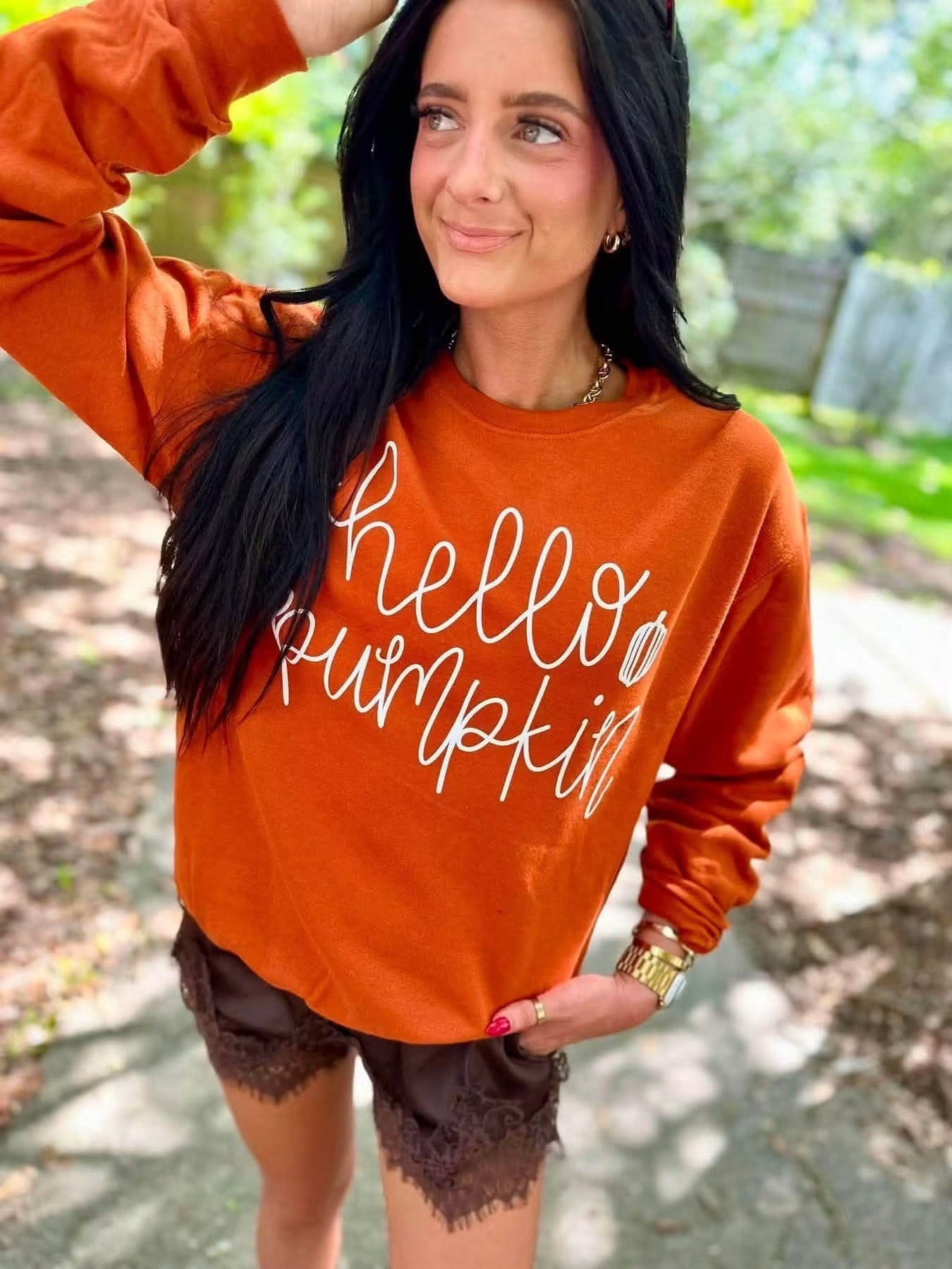A woman wearing a burnt orange crewneck sweatshirt with 'hello pumpkin' printed in white script, paired with dark brown lace-up shorts, standing outdoors on a paved path with trees in the background.