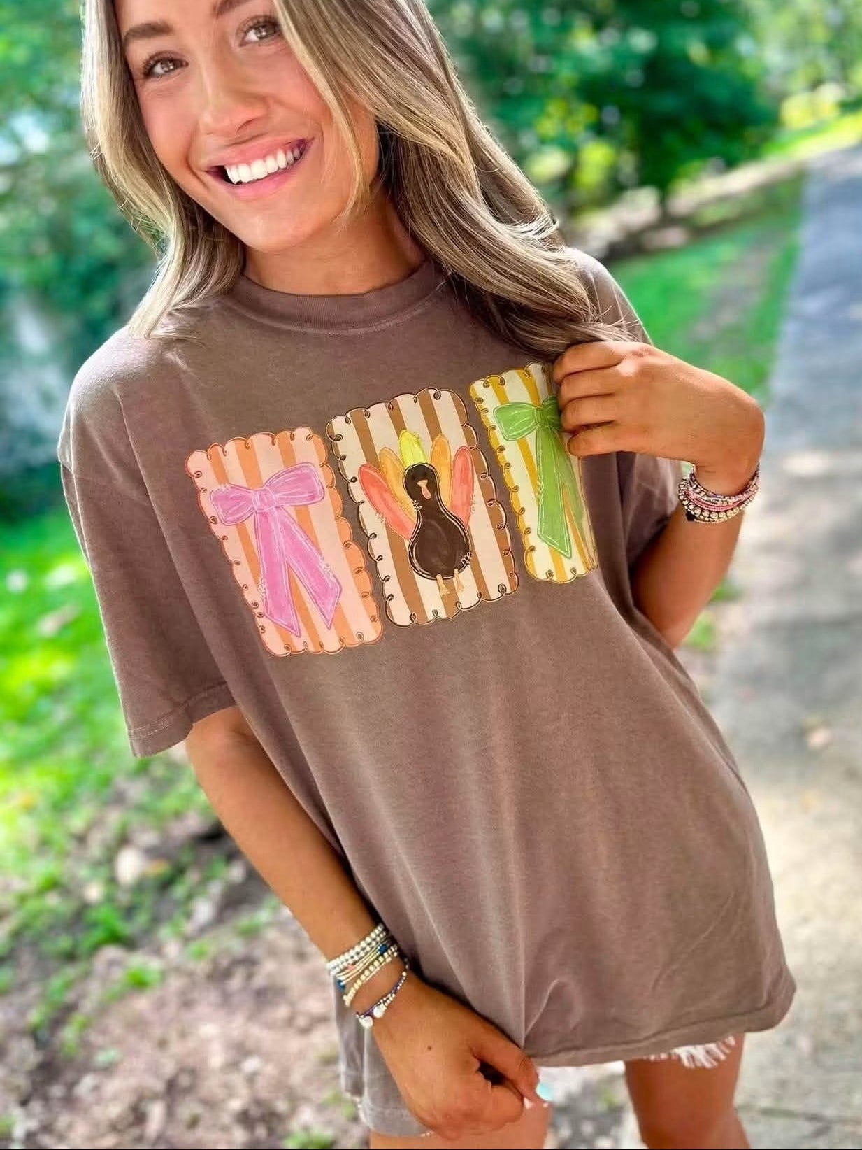 A woman wearing a brown oversized t-shirt with a colorful graphic of four striped boxes containing a pink bow, a turkey, a green corn, and a yellow flower, standing on an outdoor path.